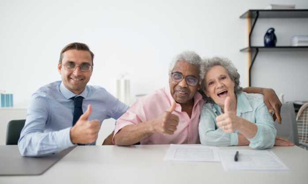 Blog - Older couple Sitting Next to a Professional and They Are all Giving a Thumbs Up to the Camera While Sitting at a White Desk